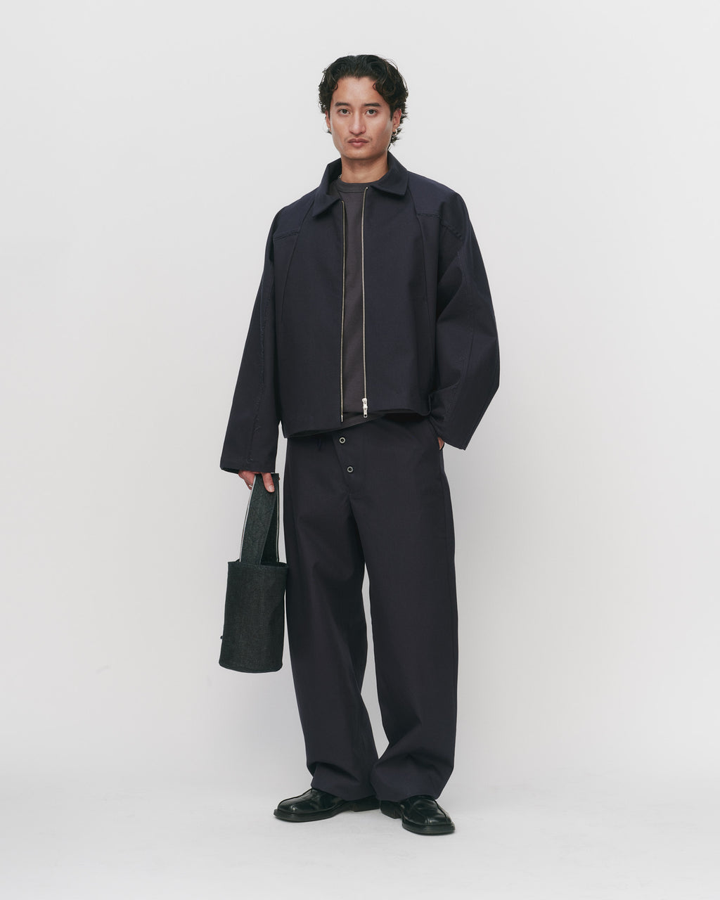 Person wearing a dark jacket and pants holding a bag on a white background