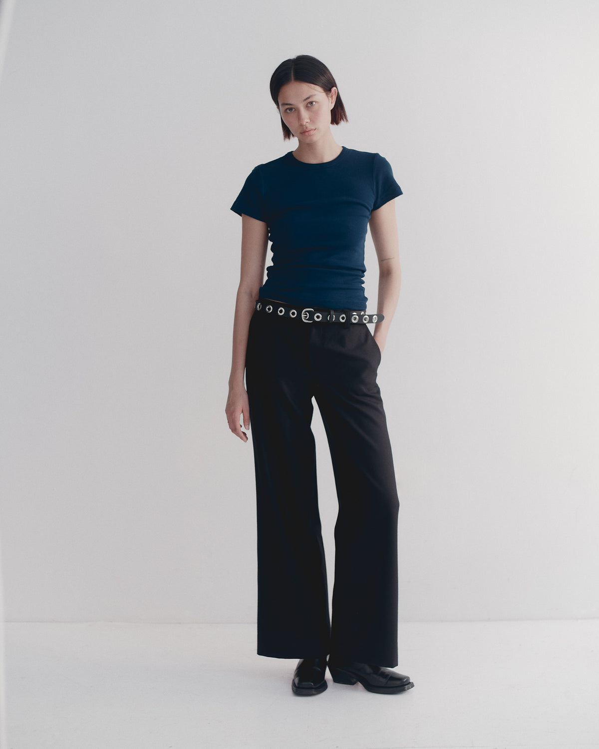 Person wearing a dark blue t-shirt and black wool tailored pants with a belt with metal rings on a white background